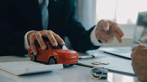 Business professionals discuss a red toy car on a desk beside documents and a key, suggesting automotive finance or a vehicle purchase.