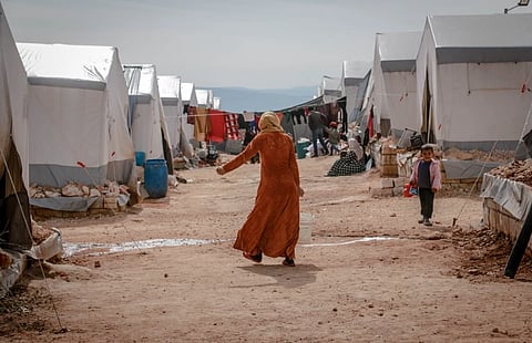 A woman in a rust-colored robe walks through a muddy refugee camp, lined with white tents, as children and families move around her.