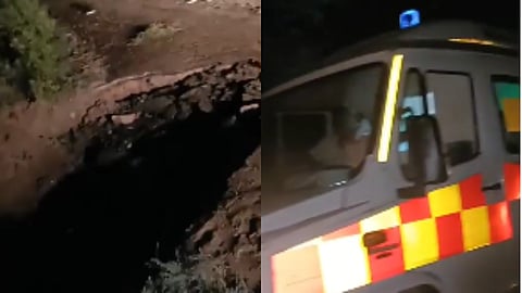 Emergency response vehicle with flashing lights beside a large, dark sinkhole or collapsed roadside embankment at night, creating a tense scene.