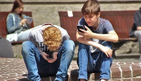 Two teenagers seated outdoors, focused on their smartphones, embracing modern technology.

