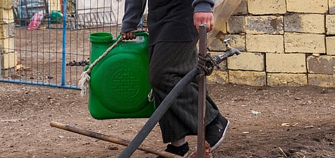 Woman carrying a green water canister next to stone wall in a rural setting, outdoors.
