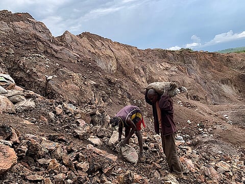 Two miners work in a barren open-pit quarry, sifting rocky earth and carrying heavy sacks under cloudy skies, conveying hardship.