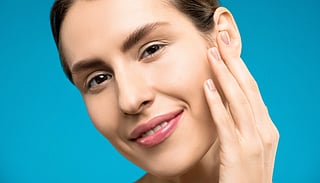 Close-up of a woman smiling with natural makeup on a blue background, showcasing beauty and skincare.
