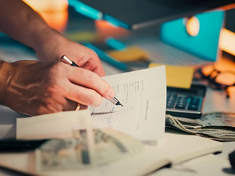 A hand signing financial documents on a desk with cash and a calculator, signifying finance management.

