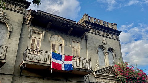 Historic ornate stone building with a Dominican flag on its balcony, framed by bougainvillea and leafy branches beneath a bright blue sky.