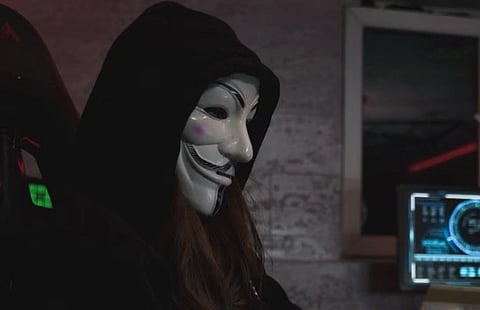 Hooded figure wearing a white Guy Fawkes mask sits beside a glowing computer monitor in a dim room, creating a secretive, ominous mood.