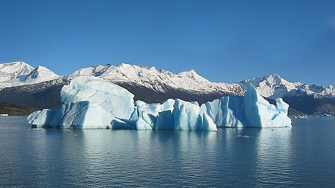Icebergs drift in the ocean, set against a backdrop of rugged mountains under a clear, sunny sky