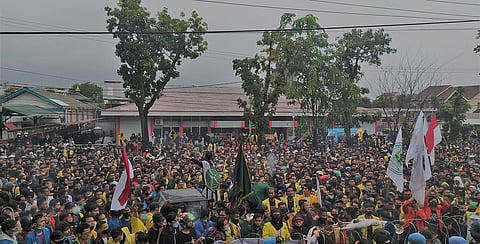 A dense outdoor crowd gathers for a street rally, waving Indonesian flags and banners beneath trees and power lines in an energetic, festive scene.