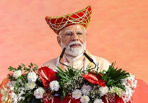Image of PM addressing at the Mahila Sashaktikaran Mahasammelan Programme held on the occasion of 300th birth anniversary of Lokmata Devi Ahilya Bai Holkar at Bhopal, in Madhya Pradesh. He is standing in front of a podium decorated with flowers and he is also wearing a turban type hat on his head.