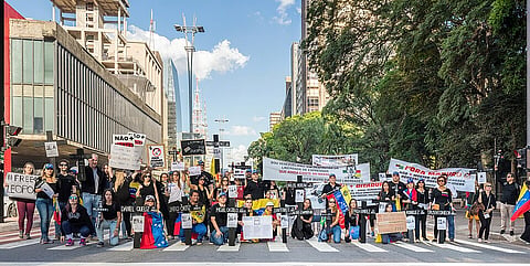 A diverse crowd of protesters fills a city street with signs and banners, beneath skyscrapers and leafy trees, in a peaceful demonstration.