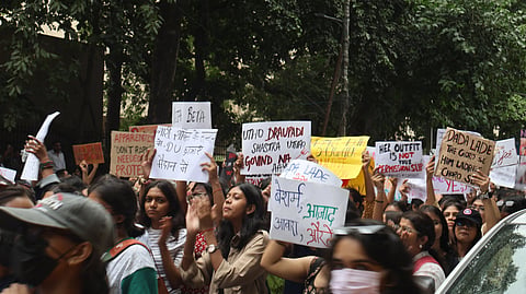 A dense protest crowd gathers beneath leafy trees, raising handwritten Hindi and English placards; determined faces convey urgency and solidarity in the women protest of Delhi university 