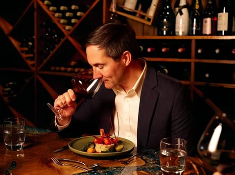 In the image a man is having luxurious food in a fine diner and is seen ravishing a glass of wine in his hand. 