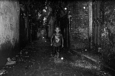 A young child joyfully runs through a dark, rainy urban alley, captured in dramatic black and white.
