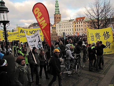 A crowded climate protest fills a European square, with marchers carrying yellow banners and signs beneath historic buildings; the mood is urgent and determined.
