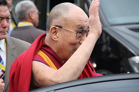 The Dalai Lama, wearing glasses and a red robe, waves from a car window as onlookers stand nearby; the scene feels warm and welcoming.