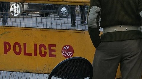 A police officer in uniform stands near another policeman sitting on a motorcycle behind a yellow barricade labeled "POLICE" on a busy street. 