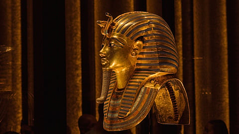 Intricately detailed Egyptian pharaoh mask on museum exhibit display with golden lighting.
