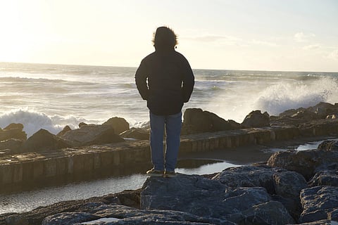 A person in a winter coat stands on rocky coastline watching ocean waves crash at sunset.
