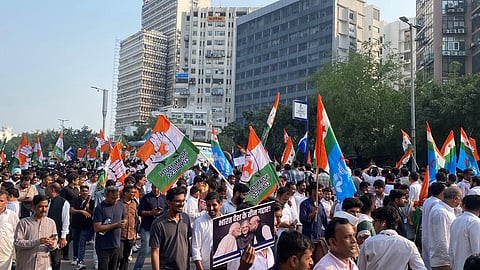 A large crowd marches through Delhi street, from Barakhamba road to the Supreme court, waving colorful political party flags, placards and satirical posters of leader; the mood is energetic and protest-like.