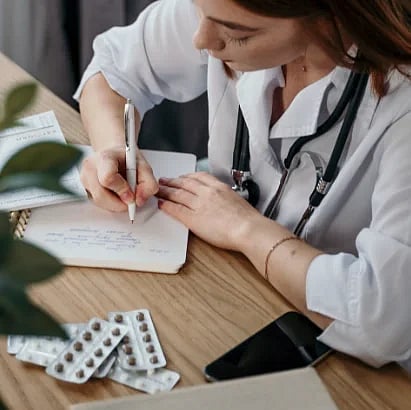 Young female doctor writing a prescription in a medical office with a stethoscope and pills nearby.
