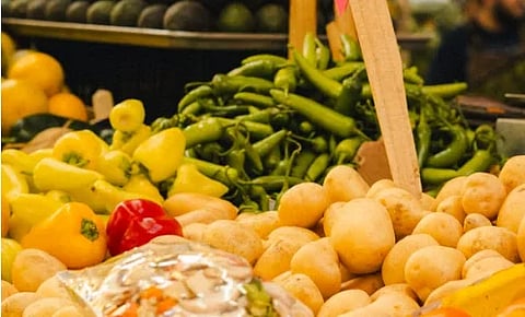 A colorful market stall displays heaps of tan potatoes, with yellow and red peppers and long green chilies behind, evoking fresh, abundant produce.