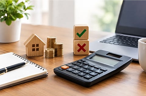 A desk calculator, model house, stacked coins, and check and X blocks sit beside a laptop and notepad, suggesting a thoughtful home-buying budget decision.