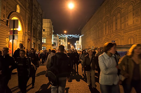 A crowd of people walking down Moscow street at night, illuminated by overhead festive lights and street lamps.


