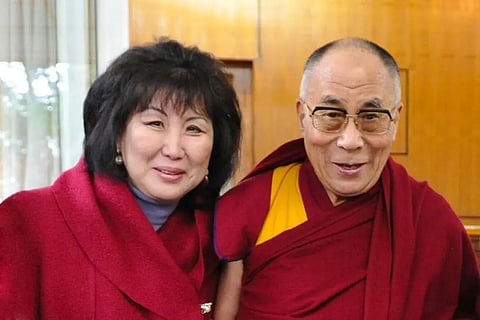 Smiling woman in a red coat stands beside the smiling Dalai Lama, in maroon robes and glasses.