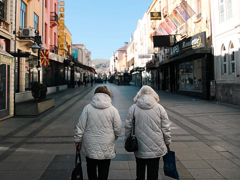 Two women in winter coats walking down a colorful city street on a clear day.
