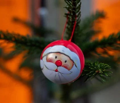 Close-up of a Santa-themed ornament hanging on a Christmas tree branch outdoors.
