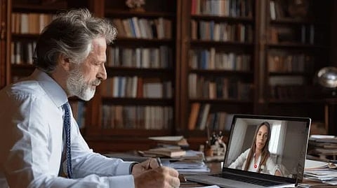 An older man reviews paperwork in a book-lined office while video-calling a woman on his laptop, creating a focused, professional mood.