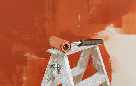 Paint roller on a ladder with a partially painted red wall in the background.
