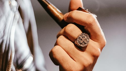 Detailed view of a hand in a leather jacket holding a pipe, wearing a vintage signet ring.
