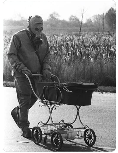 A man wearing a gas mask pushes a baby carriage down a street,