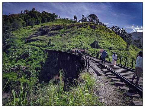 A narrow railway bridge crosses a lush green hillside as hikers walk along the tracks beneath a bright blue, cloud-streaked sky.