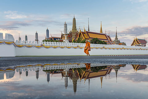A saffron-robed monk walks beside a white temple wall at Bangkok’s Grand Palace, reflected in still water beneath an expansive pastel sky.