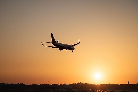 A passenger airplane descends toward the runway above a dark airport skyline, silhouetted against a glowing orange sunset; the scene feels calm and cinematic.