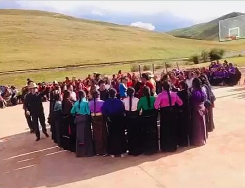 Large group in colorful traditional clothing gathers on an outdoor basketball court in a mountainous landscape.

