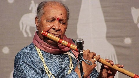 इस तस्वीर में पंडित हरिप्रसाद चौरसिया बाँसुरी बजा रहे हैं। उनके माथे पर चंदन लगा हुआ है और उन्होंने गले में माला पहन रखी है।