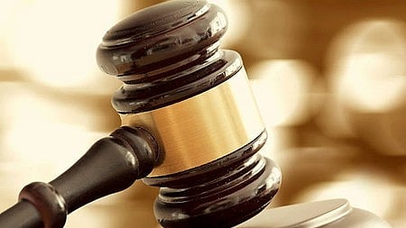 धुंधली और गर्म पृष्ठभूमि में सुनहरी पट्टी वाला गहरे रंग की लकड़ी का हथौड़ा (गैवल) अपने ब्लॉक के ऊपर रखा है। यह दृश्य न्याय या नीलामी को दर्शाता है।