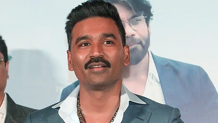 अभिनेता और निर्देशक धनुष  खड़े है मुस्कुरा रहे है 