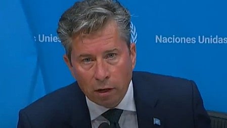 टॉम फ्लेचर बैठे है कुर्सी पर और वह वाइट और ब्लैक कलर का शर्ट और कोर्ट पहने है  