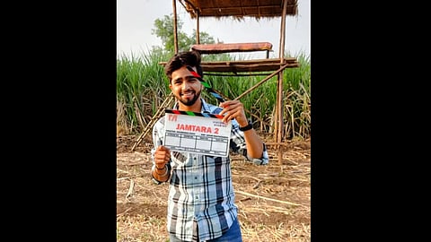 तस्वीर में एक आदमी निर्देशक के पास खड़ा है, हाथ में एक एक्शन प्रॉप लिए हुए, पीछे खेत दिखाई दे रहे हैं।