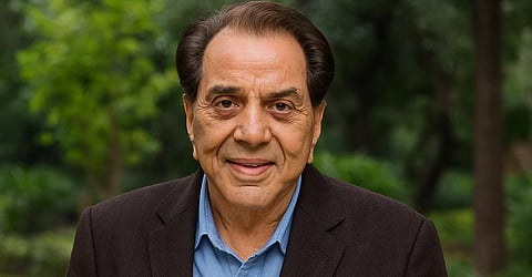 तस्वीर में धर्मेंद्र काले रंग की कोट और नीले रंग की शर्ट पहने हुए हैं।