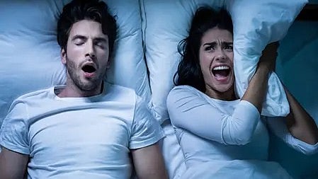 दो लोग सोते हुए, एक लड़का खर्राटे ले रहा है और लड़की कान पर तकिया रख कर परेशान है।