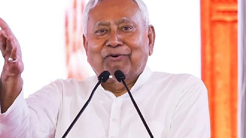 बिहार के मुख्यमंत्री नीतीश कुमार मंच पर भाषण देते हुए, सफेद कुर्ता पहने माइक के सामने हाथ उठाकर इशारा करते नजर आ रहे हैं।
