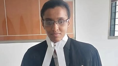 काले वकीली कोट और सफेद नेकबैंड (एडवोकेट बैंड) पहने चश्मा लगाई एक महिला, जो कमरे के अंदर खड़ी हैं और कैमरे की ओर देख रही हैं।