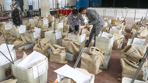 मतदान केंद्र के अंदर कर्मचारी बैलेट बॉक्स और थैलों को व्यवस्थित करते हुए, चारों ओर रखी मतपेटियां और चुनाव सामग्री दिखाई दे रही है।