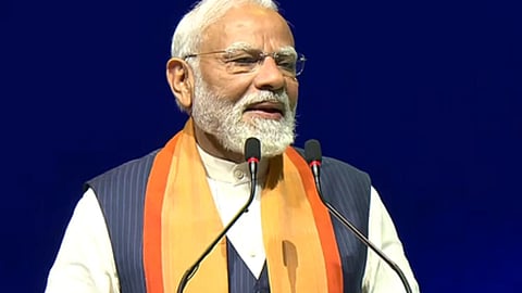 प्रधानमंत्री नरेंद्र मोदी मंच पर खड़े होकर माइक के सामने भाषण देते हुए नजर आ रहे हैं, उन्होंने सफेद कुर्ता हुआ है।