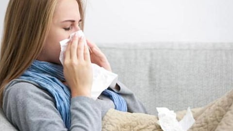 सर्दी-जुकाम से परेशान महिला सोफे पर बैठकर रुमाल से नाक साफ करती हुई।